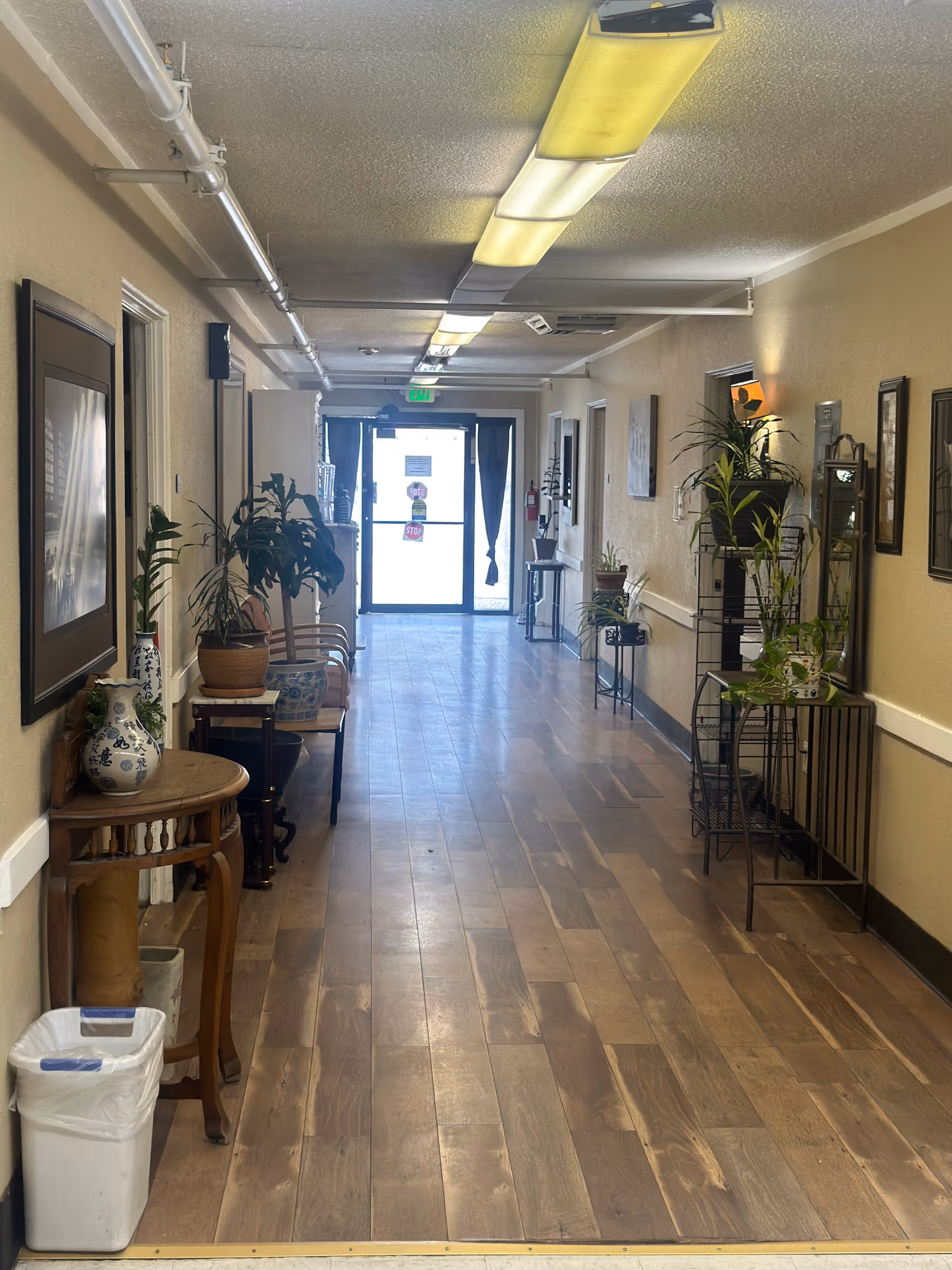 A long hallway with wooden flooring and beige walls, decorated with various potted plants on tables and stands along both sides. There are framed pictures on the walls and fluorescent lights on the ceiling. At the end of the hallway is a glass door with an exit sign above it.