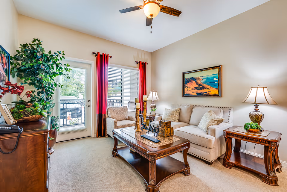 Bright furnished living room with a beige sofa, armchair, wooden coffee and side tables, decorative lamps, a ceiling fan, and a glass door with red curtains leading to a balcony.