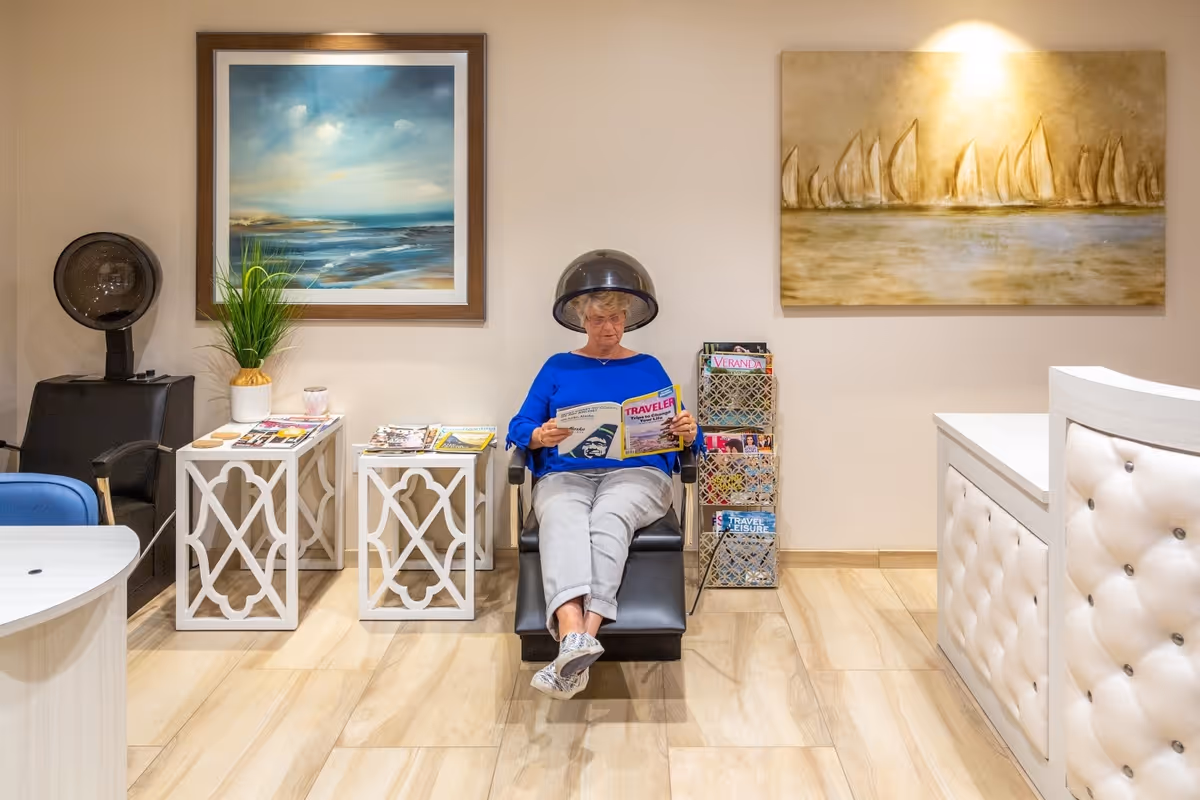 An elderly woman sitting under a hair dryer in a salon area, reading a magazine. The room has light-colored wooden flooring, two framed paintings on the wall, a plant on a small white table, and a magazine rack filled with various magazines. There is a white reception desk on the right side.