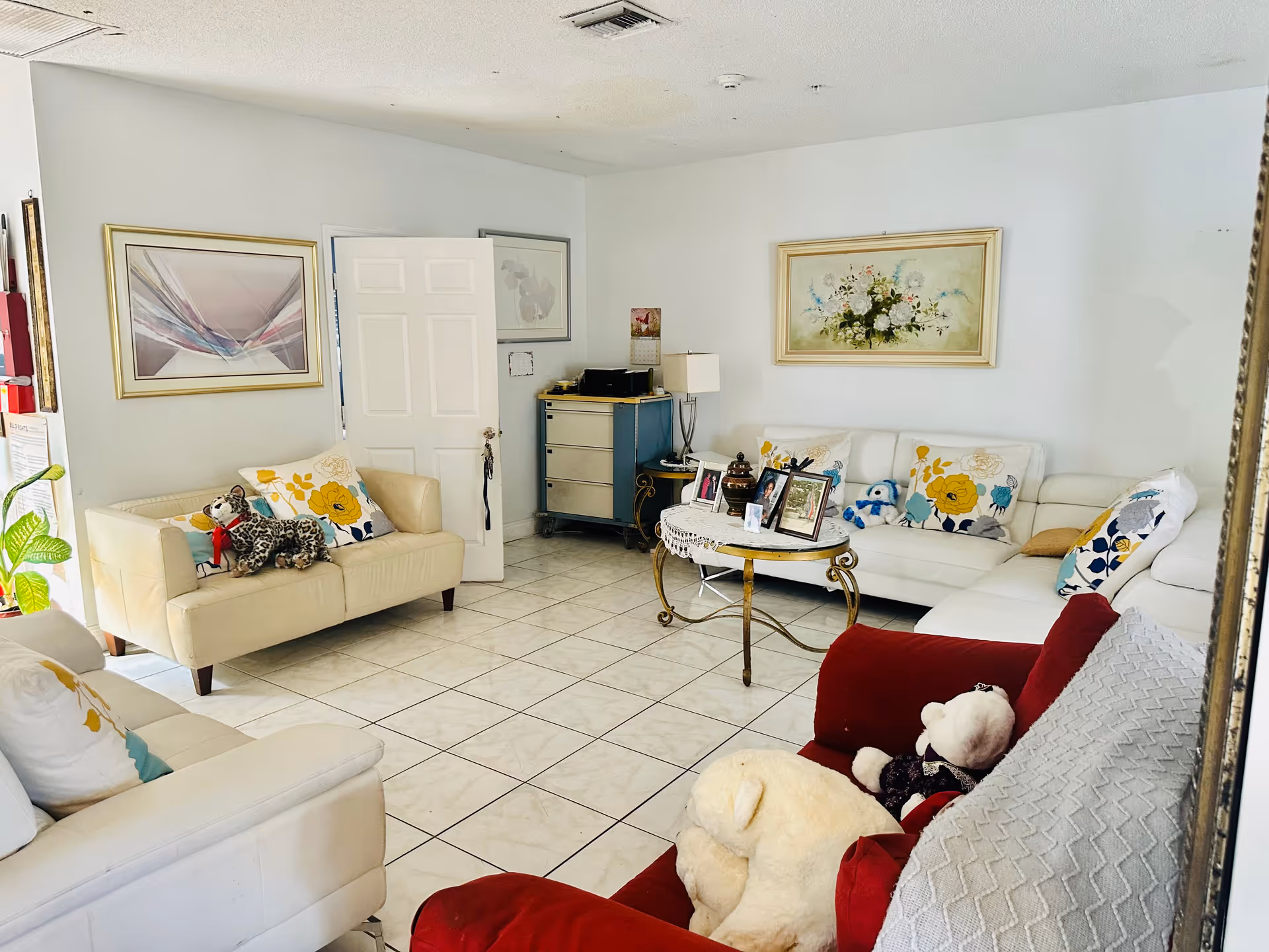 A bright living room with white tiled floor and white walls. The room contains multiple sofas with colorful floral cushions and stuffed animals placed on them. There is a glass coffee table with framed photos and decorative items in the center. The walls are decorated with framed artwork, and a small cabinet with a lamp and calendar is in the corner.