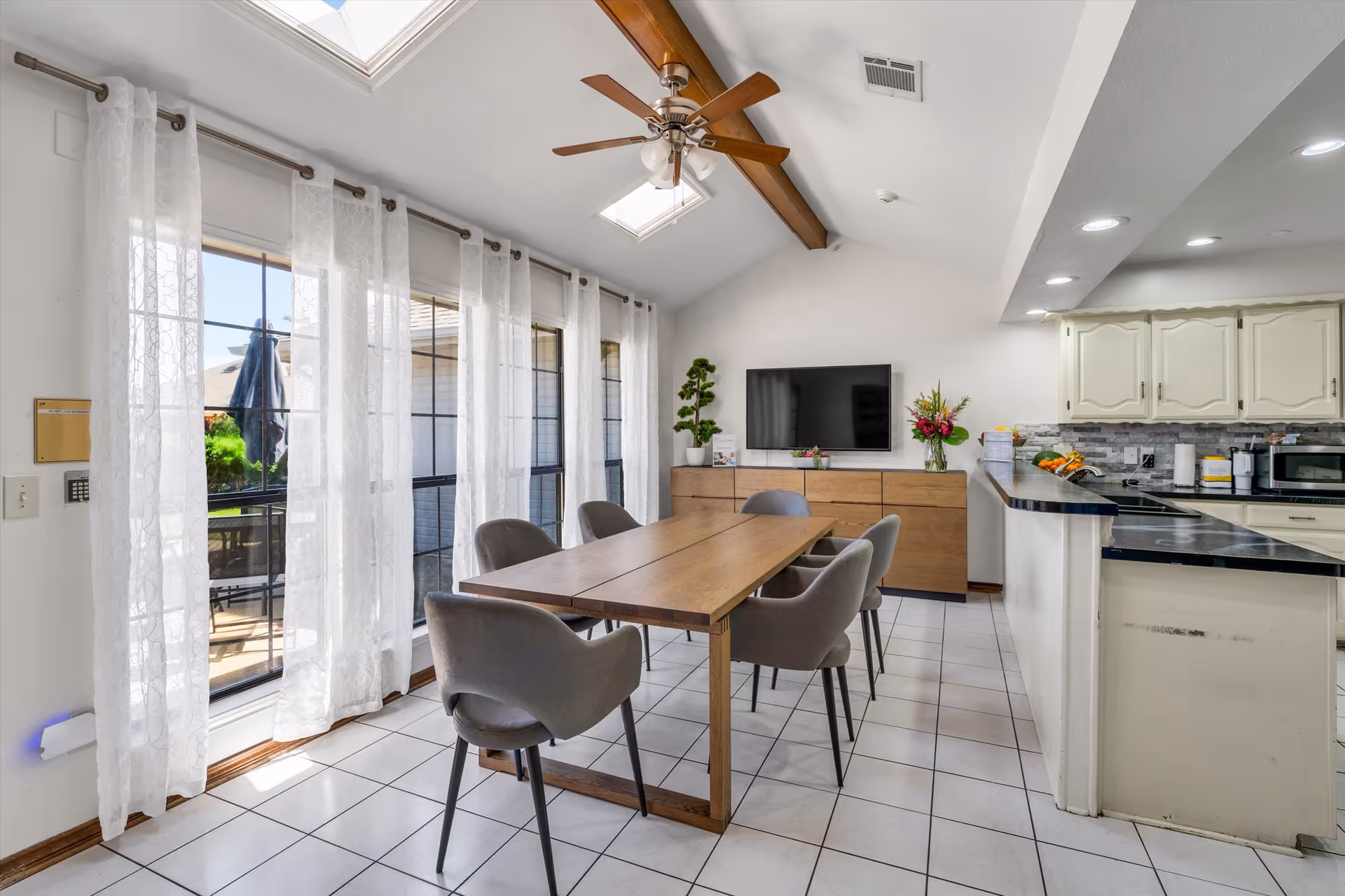 Bright dining area with a wooden table and six gray chairs, large windows with sheer curtains, a ceiling fan, and an open kitchen with cabinets and a countertop.