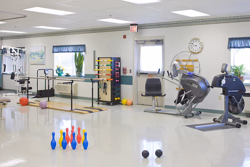 A bright senior rehabilitation exercise room with therapy equipment, parallel bars, weights, exercise bikes, and colorful plastic bowling pins on the floor.
