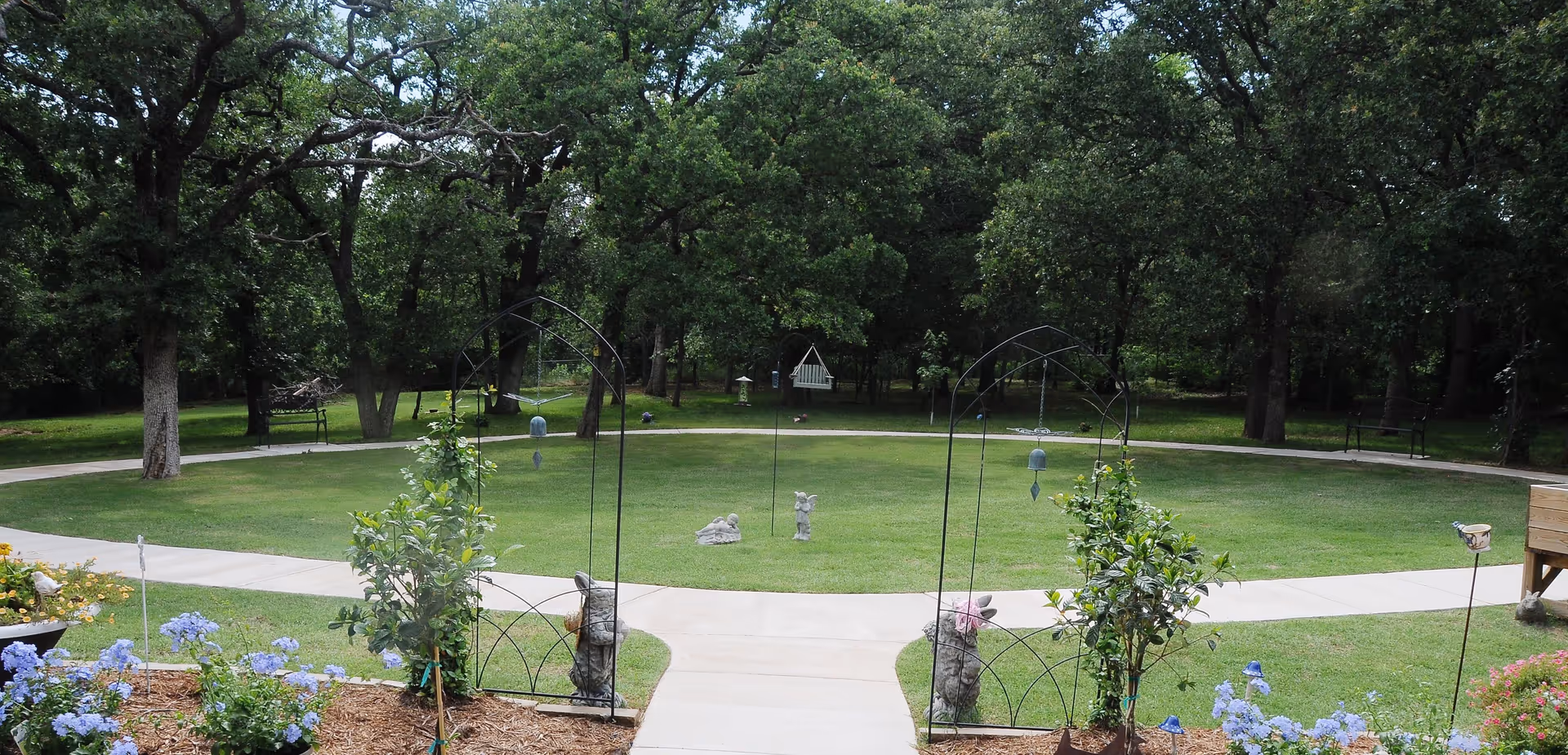 A peaceful outdoor garden area with a circular concrete pathway surrounding a grassy lawn. The garden features small statues, bird feeders, benches, and flowering plants with trees in the background providing shade.