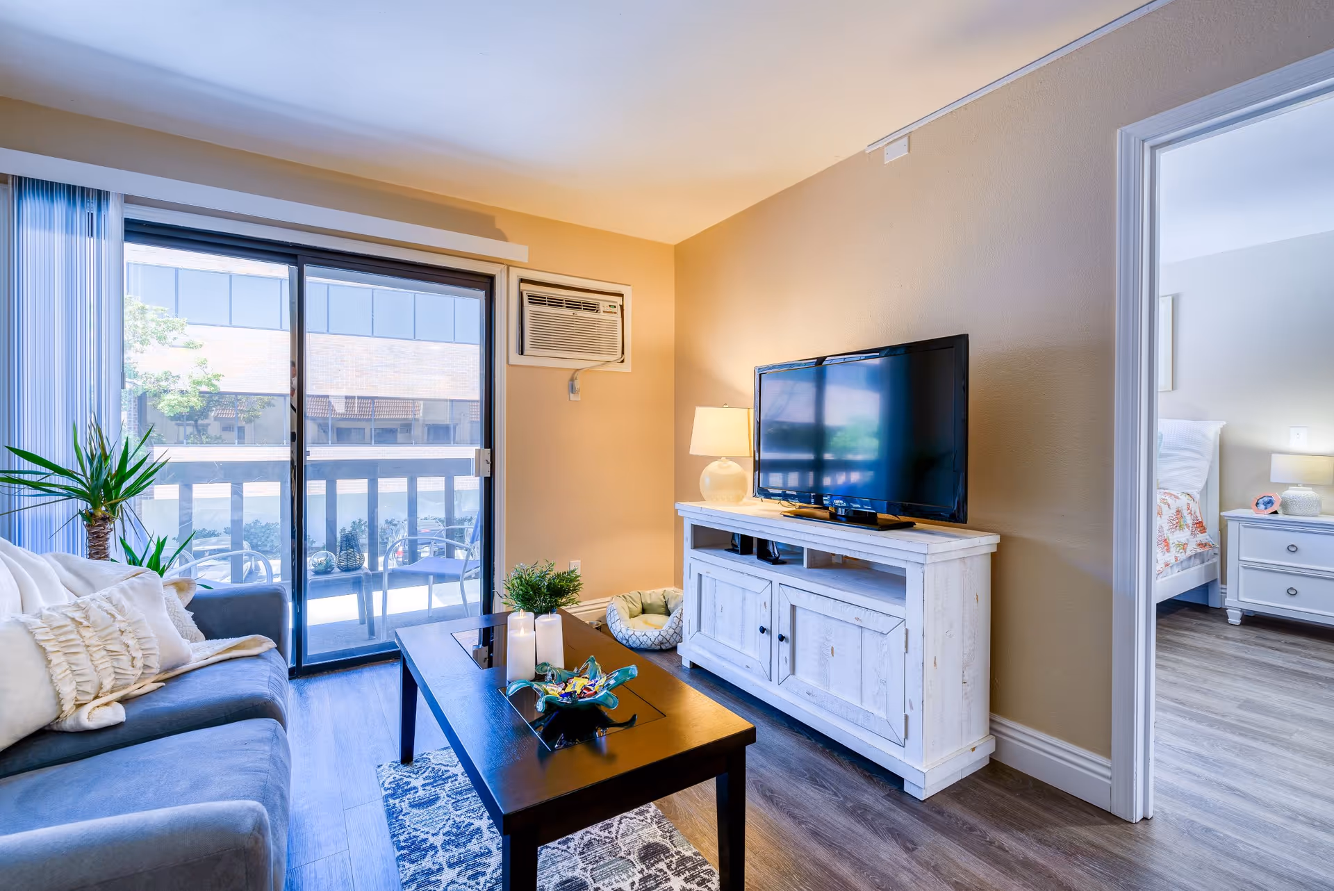 A cozy living room with a gray sofa adorned with white pillows and a throw blanket, a dark wooden coffee table with candles and decorative items, a white TV stand with a flat-screen television and a lamp, a small pet bed on the floor, and a sliding glass door leading to a balcony with outdoor chairs. The room has light beige walls and wood flooring. A doorway on the right leads to a bedroom with a white nightstand and bed.