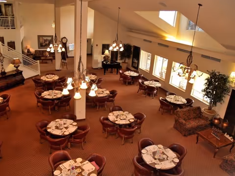 A spacious dining room with multiple round tables set for meals, each surrounded by four to six chairs. The room features warm lighting from hanging chandeliers, large windows letting in natural light, and comfortable seating areas with sofas and a coffee table. There is a grand piano in the corner and decorative plants throughout the space.