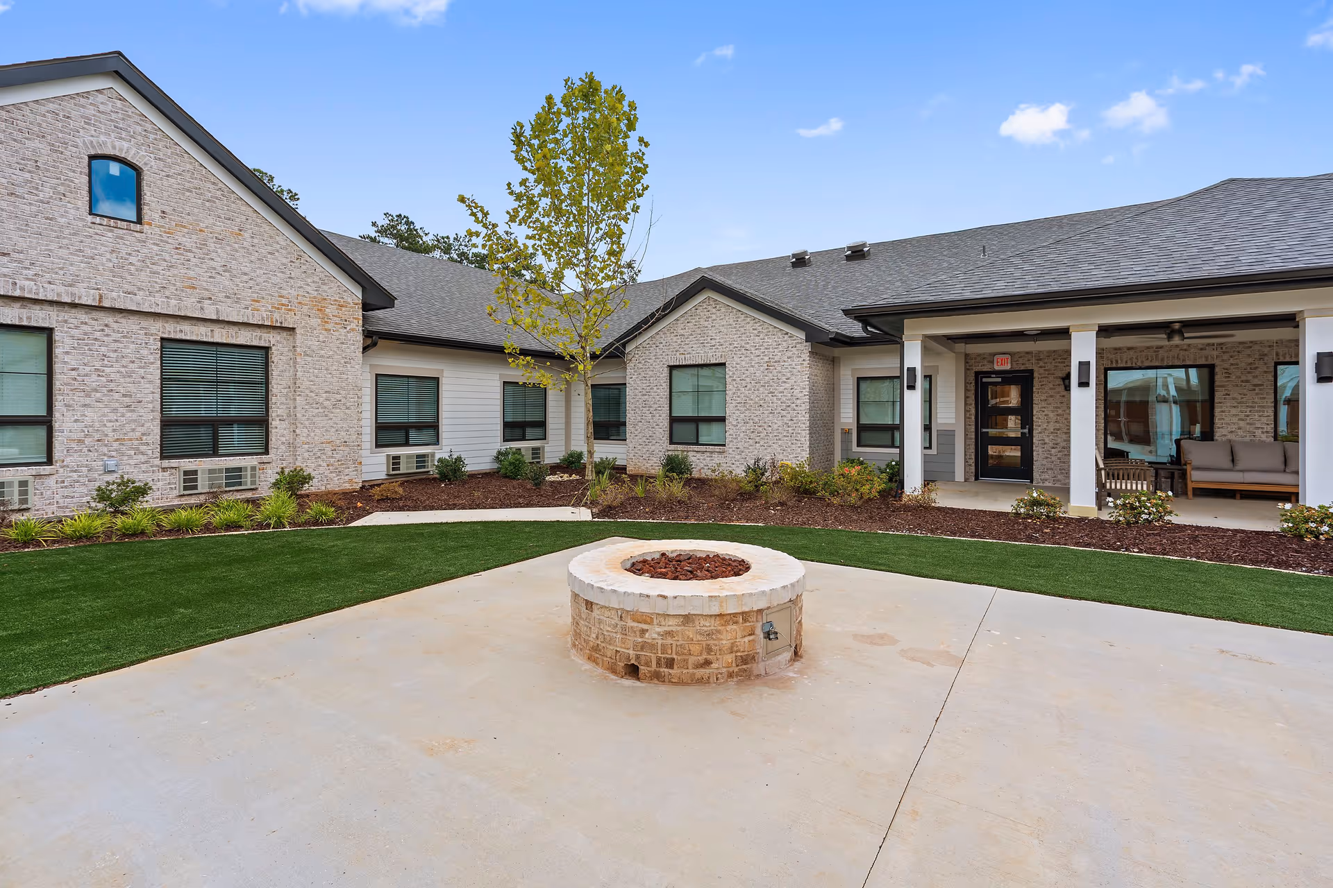 Courtyard with a central brick fire pit in front of a single-story brick building and covered patio seating.
