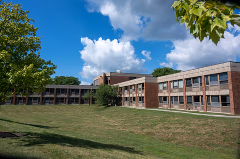 Exterior view of a two-story brick building with many windows, surrounded by a grassy lawn and trees under a partly cloudy blue sky.