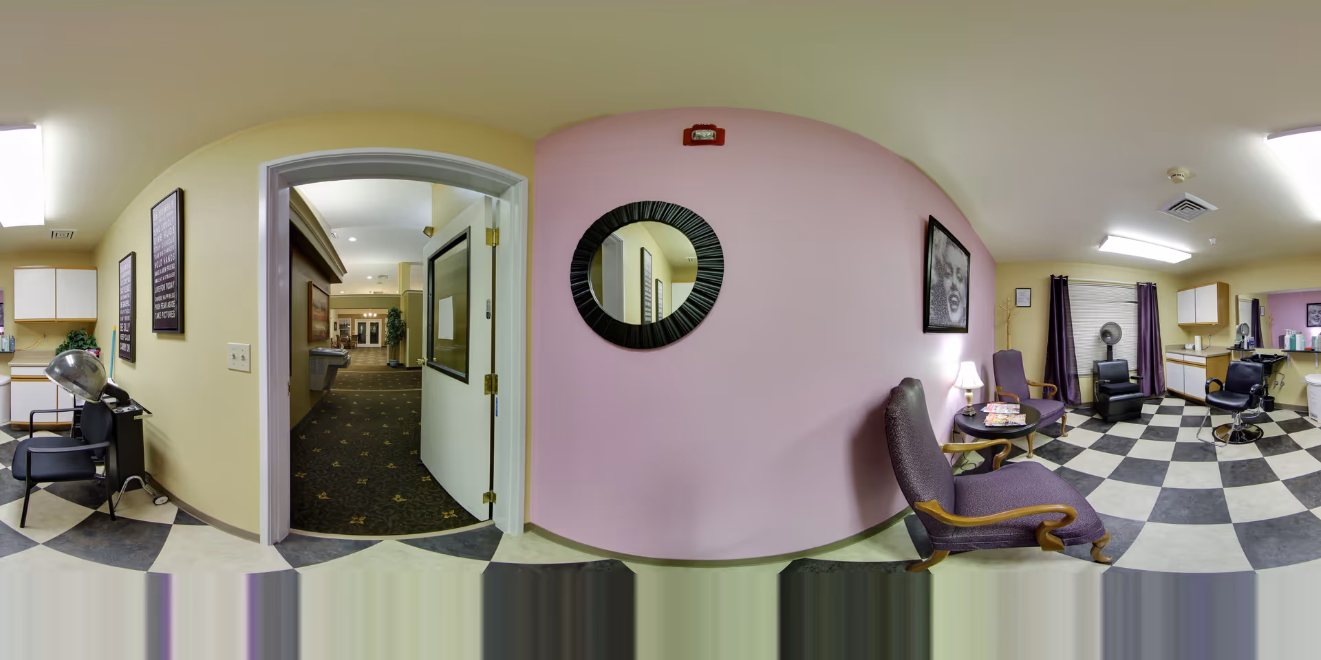 Interior view of a hair salon area in a senior living facility with checkered black and white flooring, purple walls, and several salon chairs. There is a round mirror on the wall, framed artwork, a small table with a lamp, and a hallway visible through an open door.