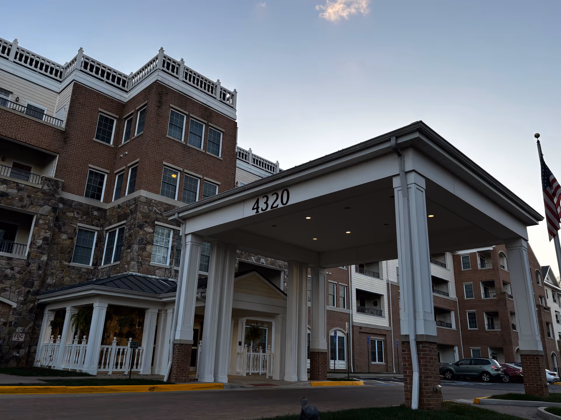 Front entrance and covered portico of a multi-story brick senior living building marked 4320 with an American flag at the curb.