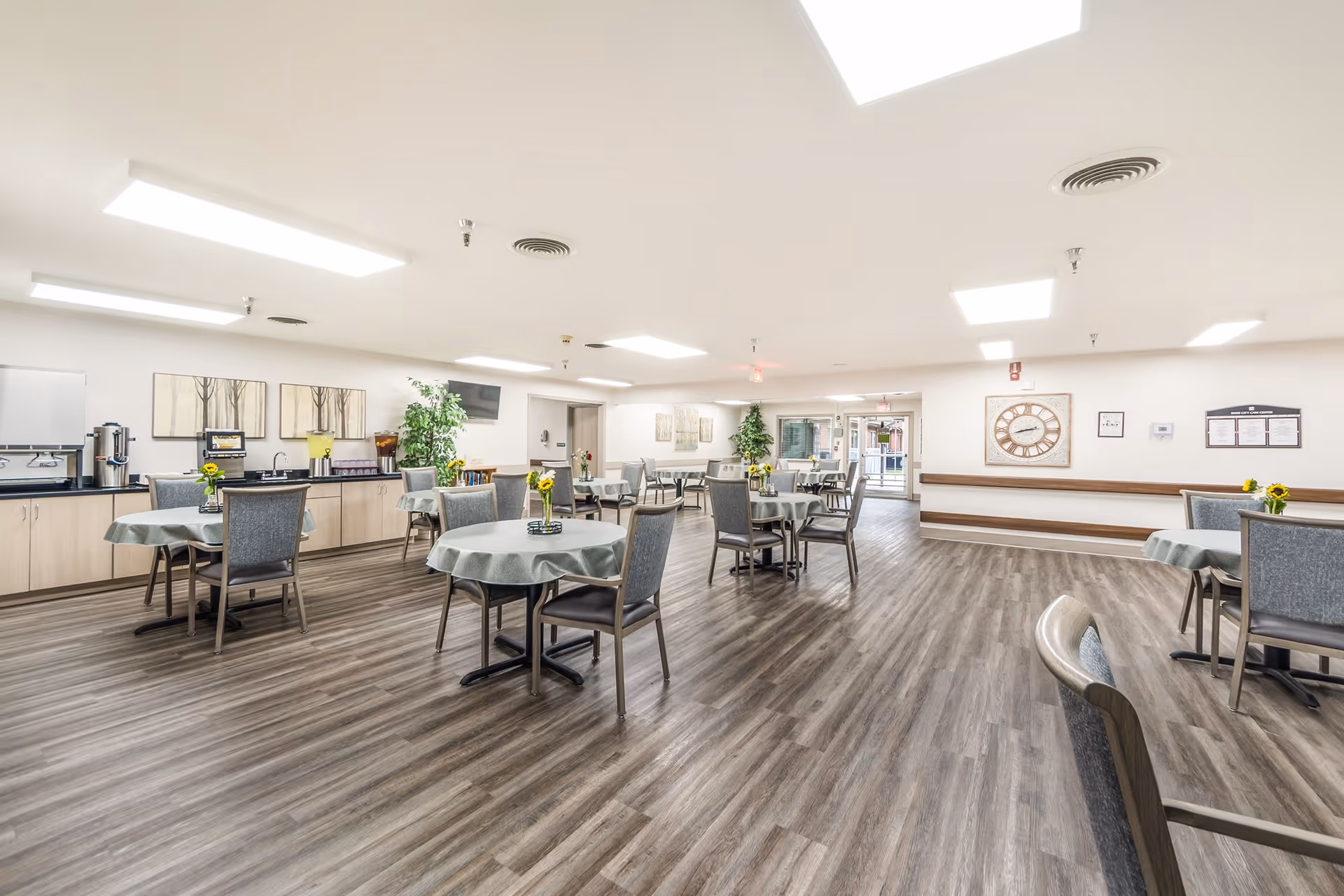 Spacious senior living dining room with round tables and chairs, wood-look flooring, and a service counter along the wall.