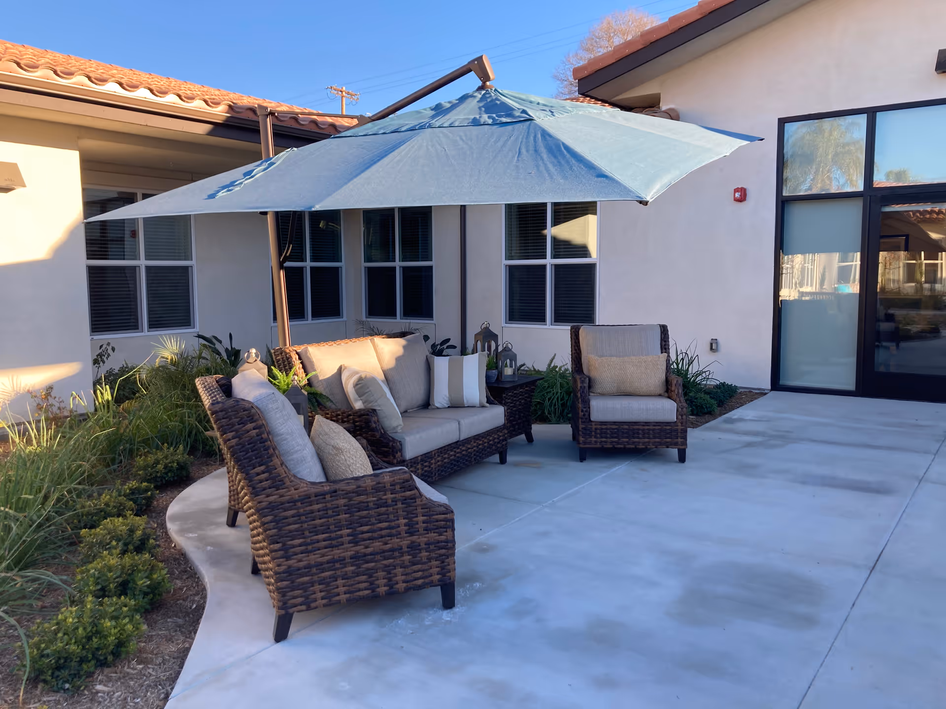 Outdoor patio area with wicker furniture including a sofa and two armchairs with cushions and pillows, shaded by a large blue umbrella. The patio is adjacent to a building with windows and a glass door, surrounded by some greenery and plants.