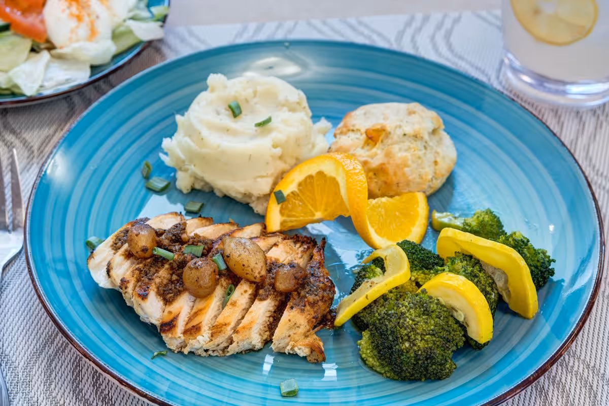 A blue plate with a meal consisting of grilled chicken topped with small roasted potatoes and chopped green onions, mashed potatoes garnished with green onions, a biscuit, steamed broccoli with yellow squash slices, and two orange slices. A glass of water with lemon and a side salad are partially visible on the table.