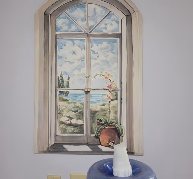 Mural of an arched window showing a seaside landscape with a potted orchid on the sill and stacked disposable cups on a water cooler below.