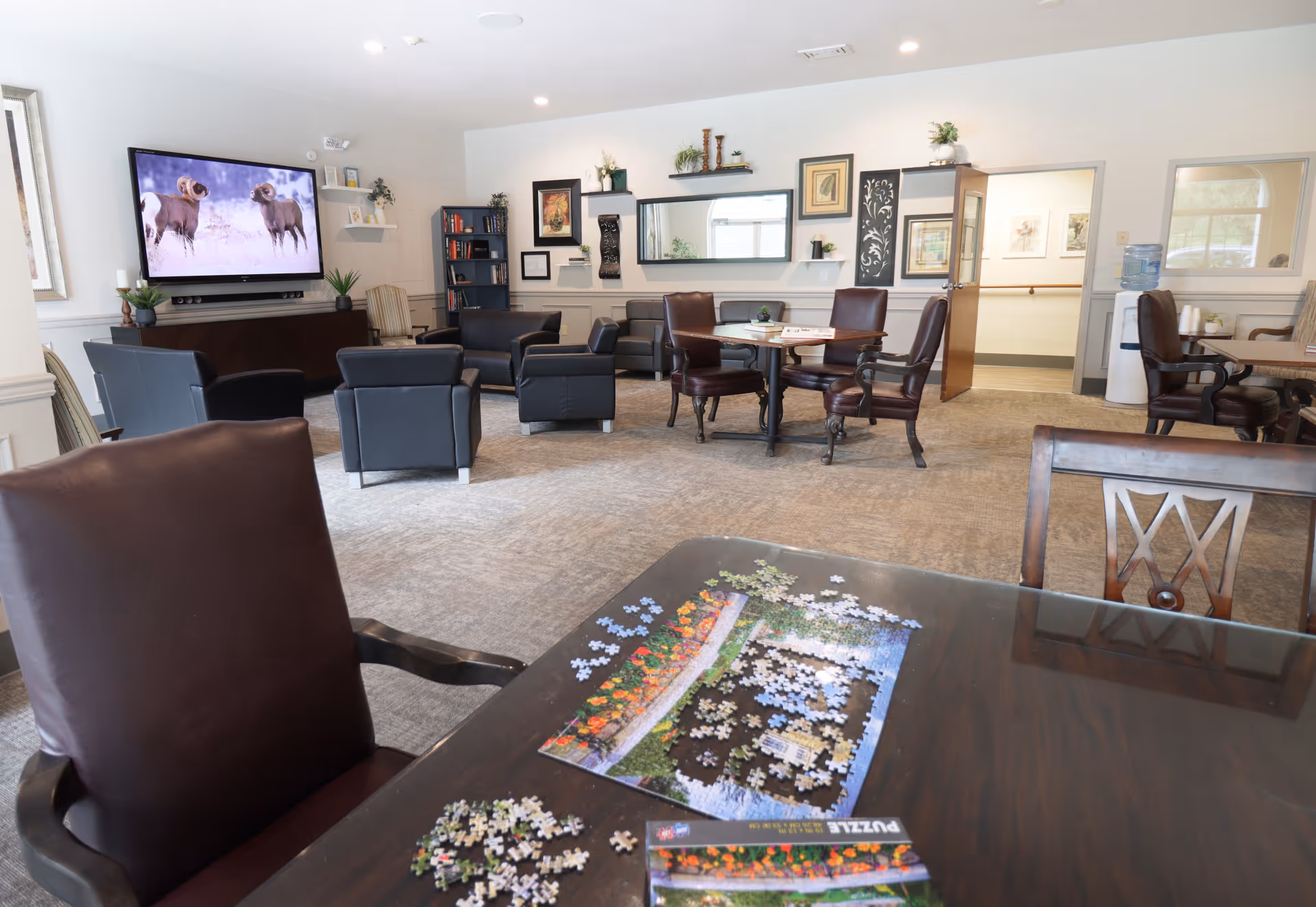 A spacious common area in a senior living facility with several seating arrangements including leather chairs and a table with a partially completed jigsaw puzzle. A large flat-screen TV on the wall displays an image of two rams. The room is decorated with framed artwork, plants, and shelves, and has a carpeted floor and bright lighting.