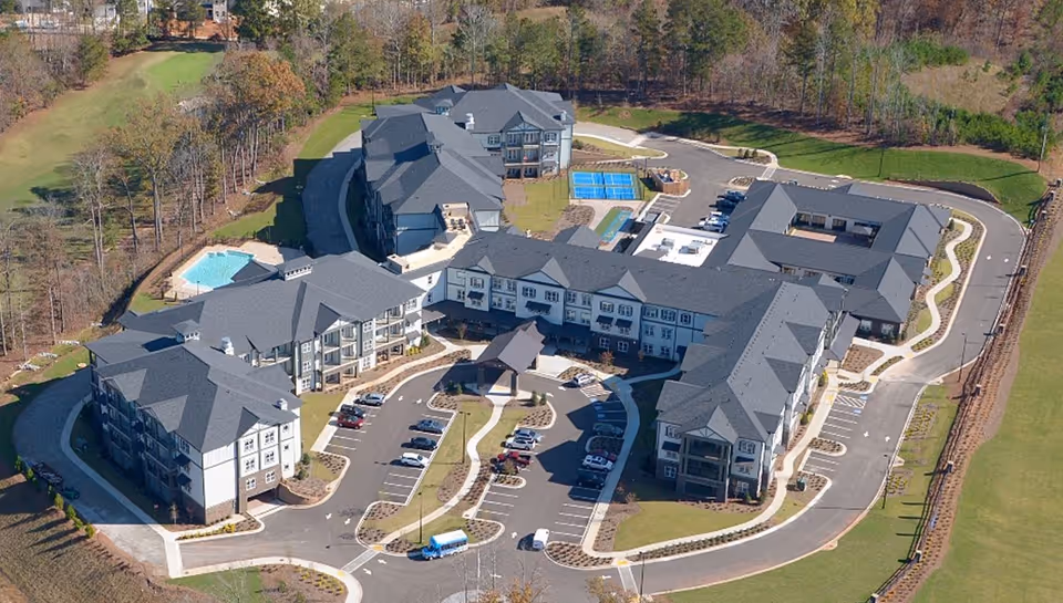 Aerial view of The Phoenix at Braselton senior living facility showing multiple connected buildings with gray roofs, parking lots with cars, a swimming pool, tennis courts, and surrounding wooded areas.