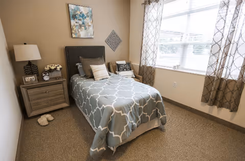A small, neatly arranged bedroom with a single bed covered in a blue and white patterned comforter. The bed has multiple pillows and a dark upholstered headboard. Next to the bed is a wooden nightstand with a lamp, a clock, a framed photo, and a small vase of flowers. There are slippers on the carpeted floor beside the nightstand. The room has beige walls, a large window with patterned curtains, and two pieces of wall art above the bed.
