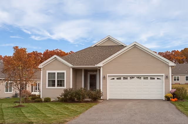 Single-story house with beige siding and a two-car garage, surrounded by a well-maintained lawn and autumn-colored trees under a partly cloudy sky.