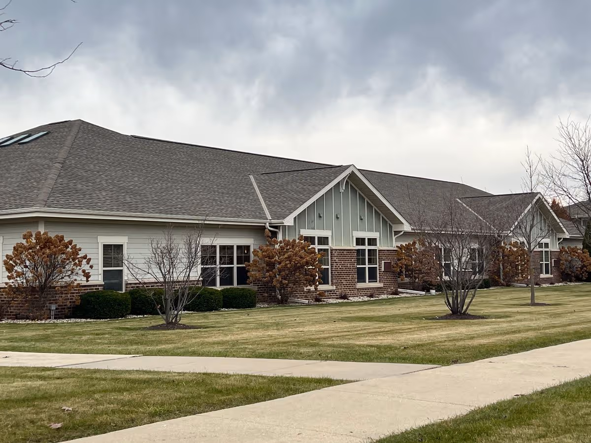Single-story brick-and-siding memory care building with a lawn, shrubs, and a sidewalk under a cloudy sky.