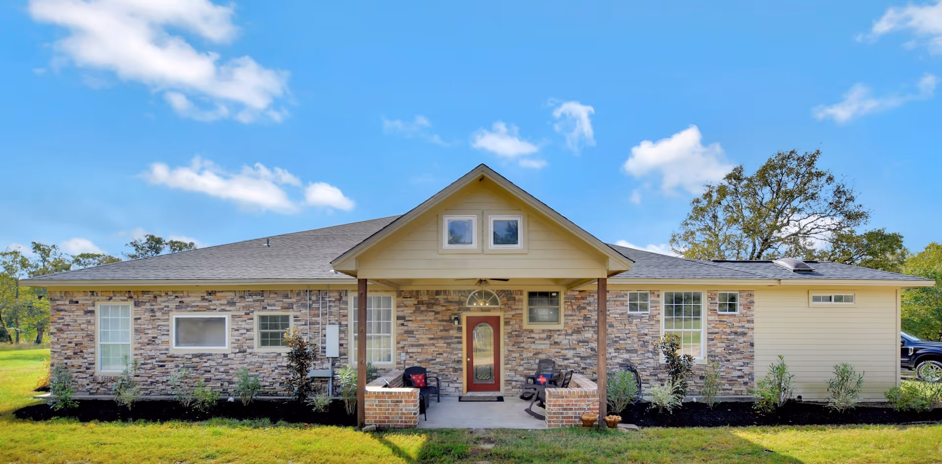 Front exterior view of a single-story assisted living facility building with a stone and beige siding facade, a covered porch with seating, and a red front door under a clear blue sky.