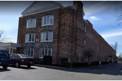 Exterior view of a multi-story brick building with several windows and a triangular roof section. There are a few parked cars in front of the building under a clear sky.