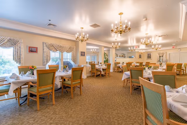 A spacious dining room with multiple tables covered in white tablecloths, each set with napkins, utensils, and small flower arrangements. The room features green cushioned wooden chairs, beige carpet, large windows with patterned curtains, and elegant chandeliers hanging from the ceiling.