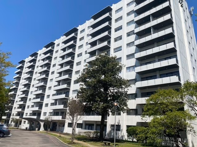 White multi-story residential building with balconies, trees, and a driveway under a clear blue sky.