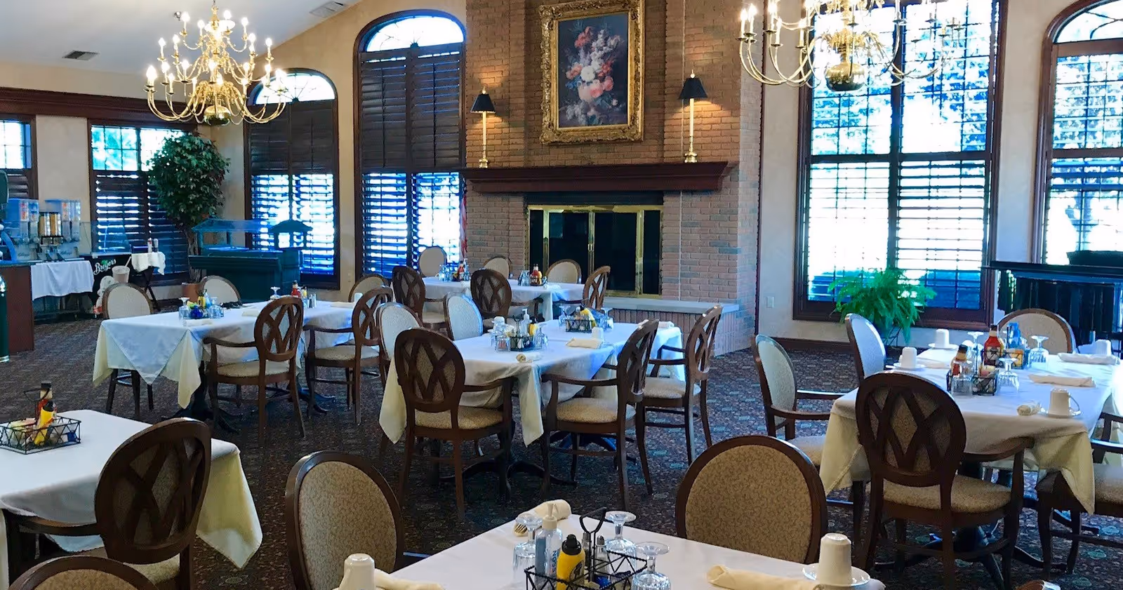 A dining room with several tables covered in white tablecloths, each set with glasses, napkins, and condiments. The room features large windows with wooden shutters, a brick fireplace with a floral painting above it, two chandeliers, and a carpeted floor. There is also a buffet area with beverage dispensers and a green plant near the windows.