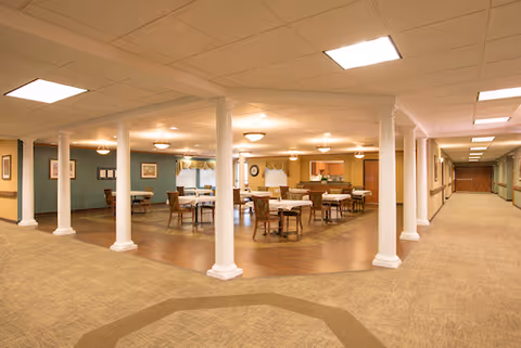 Interior view of a senior living facility dining area with multiple tables and chairs arranged on a wooden floor section, surrounded by white columns. The surrounding area has carpet flooring and a long hallway extending to the right with doors along the walls.