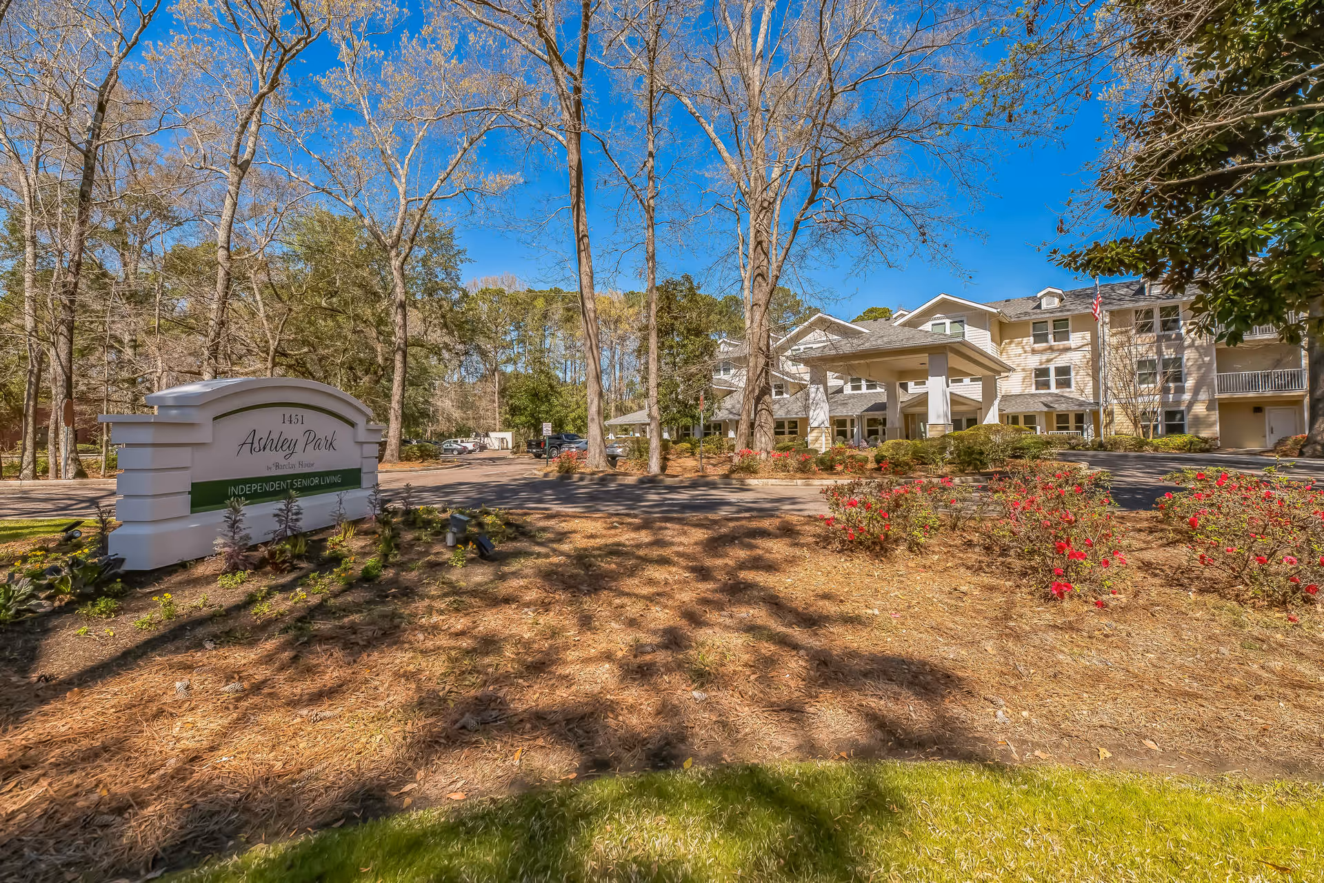 Exterior view of Ashley Park by Barclay House, an independent senior living facility, showing the main building with a covered entrance, surrounded by trees and landscaped grounds with flowers and a clear blue sky.