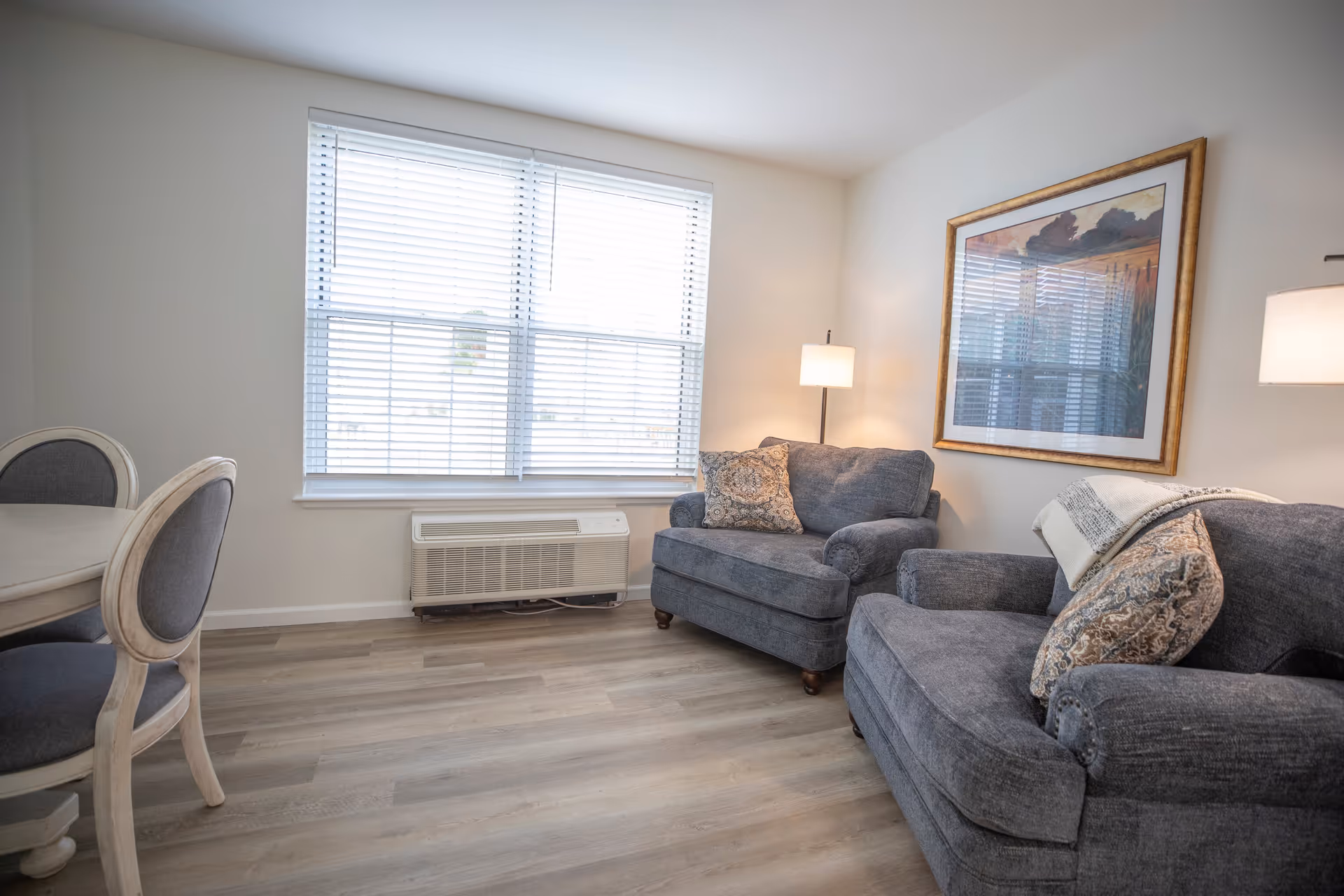 A bright living room with two gray upholstered armchairs, each with decorative pillows. A floor lamp stands between the chairs, and a large framed artwork hangs on the wall. There is a window with white blinds letting in natural light, and a white air conditioning unit below the window. Part of a dining table with gray cushioned chairs is visible on the left side. The floor is light wood.