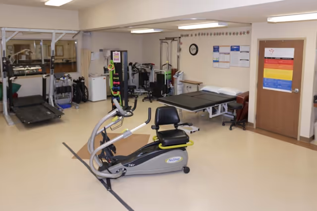 A rehabilitation room with exercise equipment including a NuStep recumbent cross trainer, a treadmill, and various physical therapy tools. There is a treatment table with pillows, a clock on the wall, and informational posters near a closed wooden door.