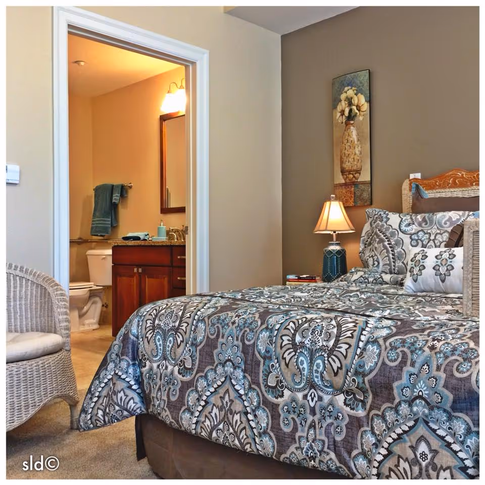 A bedroom with a patterned bedspread in shades of blue, gray, and white. There is a wicker chair with a cushion on the left side. A bedside table with a lamp and some books is next to the bed. On the wall above the bedside table is a vertical painting of a vase with flowers. Through an open doorway, a bathroom is visible with a toilet, a wooden vanity with a sink, a mirror, and a towel hanging on a rack.