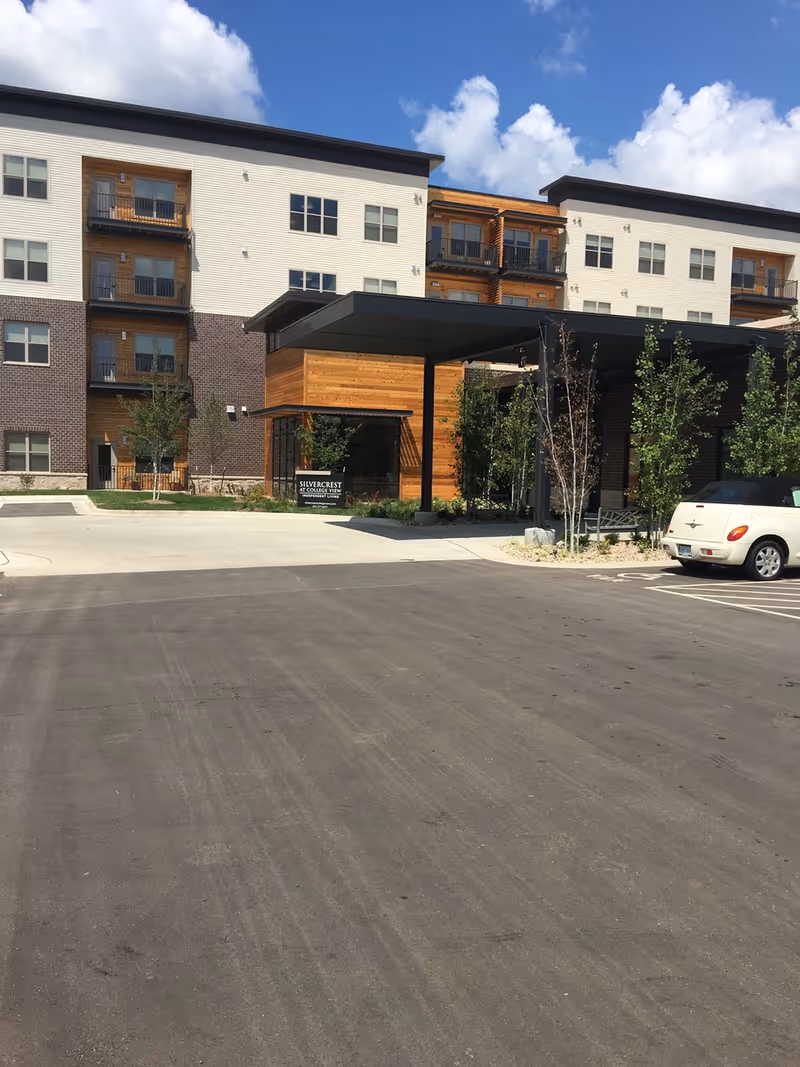 Front entrance of a multi-story senior living building with a covered drop-off, parking lot, and landscaping.