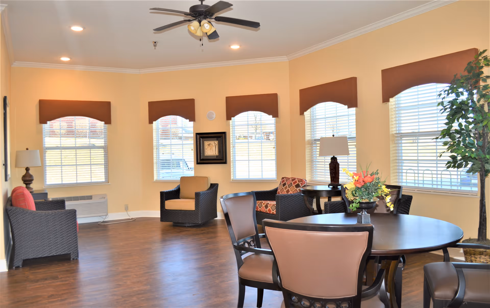 A bright and spacious common area with multiple windows covered by brown valances, several cushioned chairs, a round table with a floral centerpiece, two table lamps, and a potted plant in the corner. The room has light yellow walls and wood flooring.