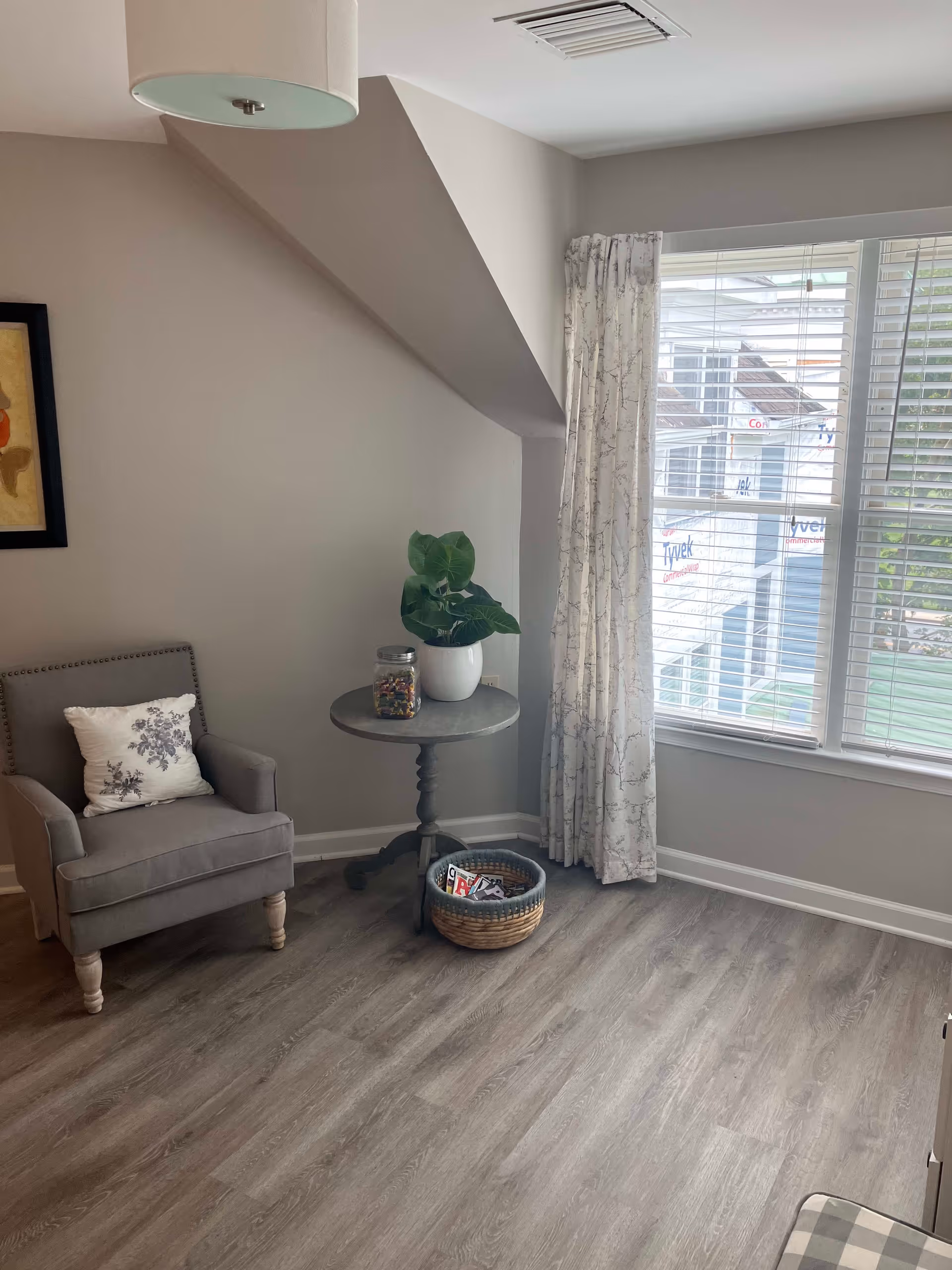 A cozy corner of a room with a gray upholstered armchair featuring a floral pillow, a small round wooden table with a potted plant and a jar on it, and a woven basket on the floor containing magazines. The room has light gray walls, wood flooring, a large window with white blinds and patterned curtains, and a ceiling light fixture.