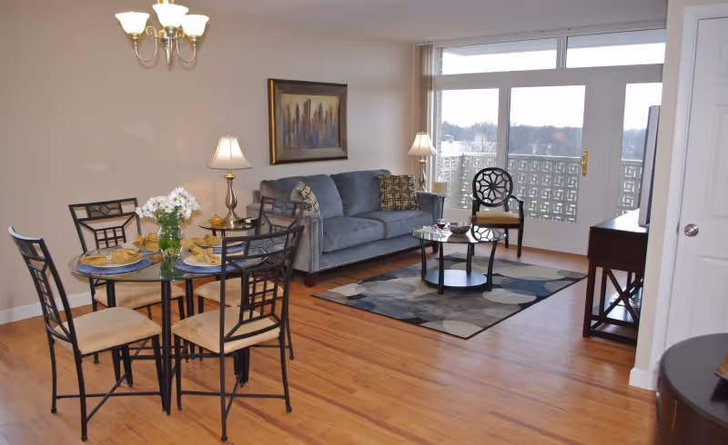 A bright living and dining area with wooden flooring. The dining area features a round glass table set with four chairs and a vase of white flowers. The living area has a gray sofa with patterned cushions, a round coffee table on a patterned rug, and a single chair near a large window with a balcony view. Two table lamps and a chandelier provide lighting, and a framed artwork hangs on the wall.