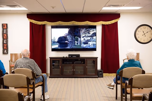 Two elderly residents sit in chairs facing a wall-mounted TV framed by red curtains in a communal TV room.