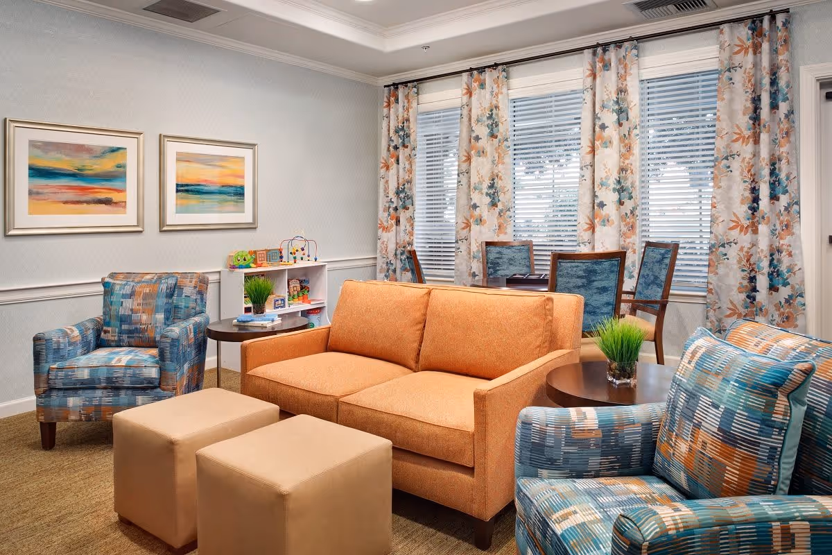 A cozy living room area with an orange sofa, two patterned armchairs, and two beige ottomans. There are two round side tables with small green plants on them. Behind the sofa, there is a small white shelf with children's toys and books. The room has large windows with floral curtains and two colorful abstract paintings on the wall.