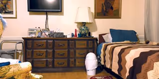 A cozy bedroom in a senior living facility featuring a wooden dresser with multiple drawers, a lamp, framed pictures, and decorative items on top. Next to the dresser is a bed with a striped brown, beige, and cream blanket and blue pillows. A walker is partially visible on the left side, and a small white trash can is on the floor near the bed. The room has framed artwork on the walls and a wooden floor.