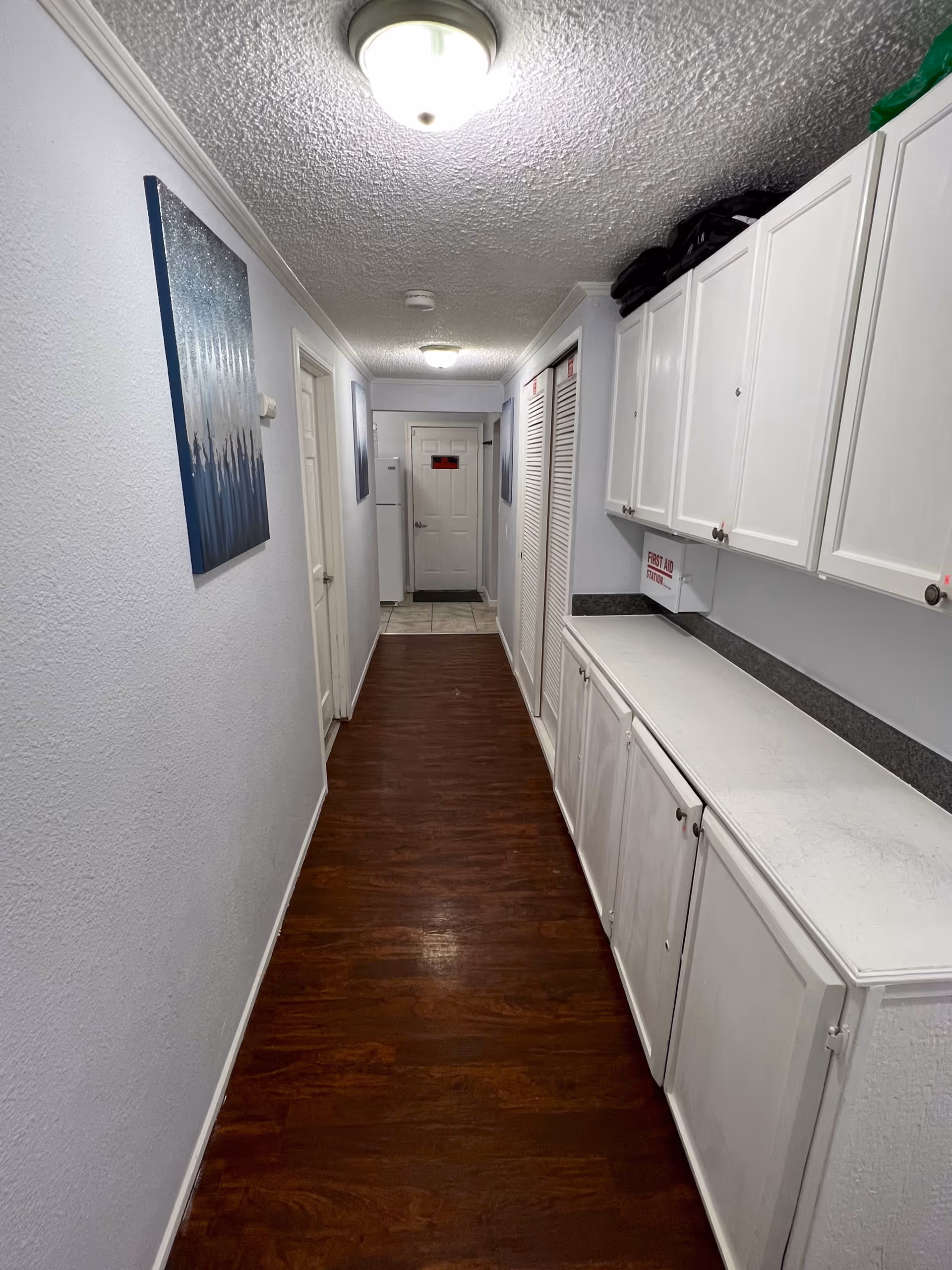 A narrow hallway with dark wooden flooring and white walls. On the right side, there are white cabinets with a countertop and a first aid station mounted on the wall. On the left side, there are two paintings hanging on the wall. At the end of the hallway, there is a white door and a refrigerator next to it. The ceiling has two round light fixtures.