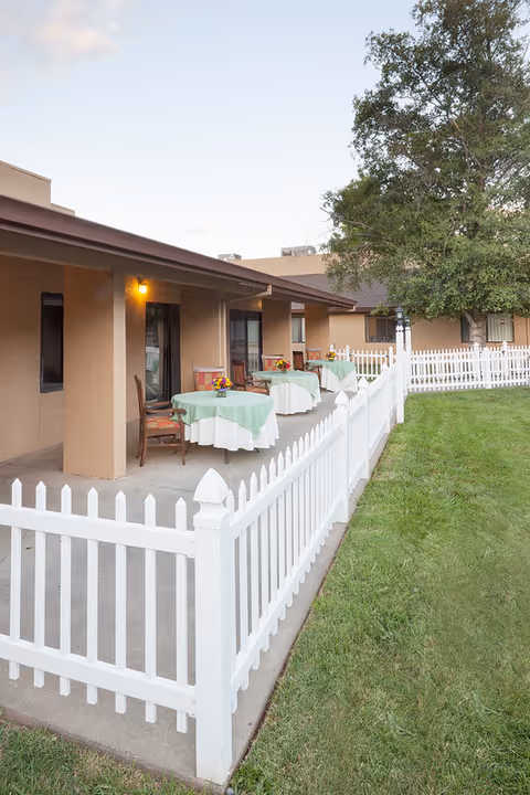 Covered patio beside a beige building with round tables draped in green-and-white tablecloths, chairs, and a white picket fence on a grassy lawn.