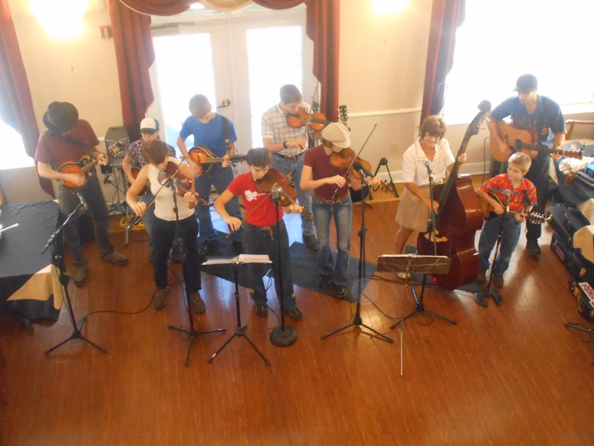 A group of people playing various string instruments including violins, guitars, a mandolin, and a double bass in a room with wooden floors and large windows with red curtains.