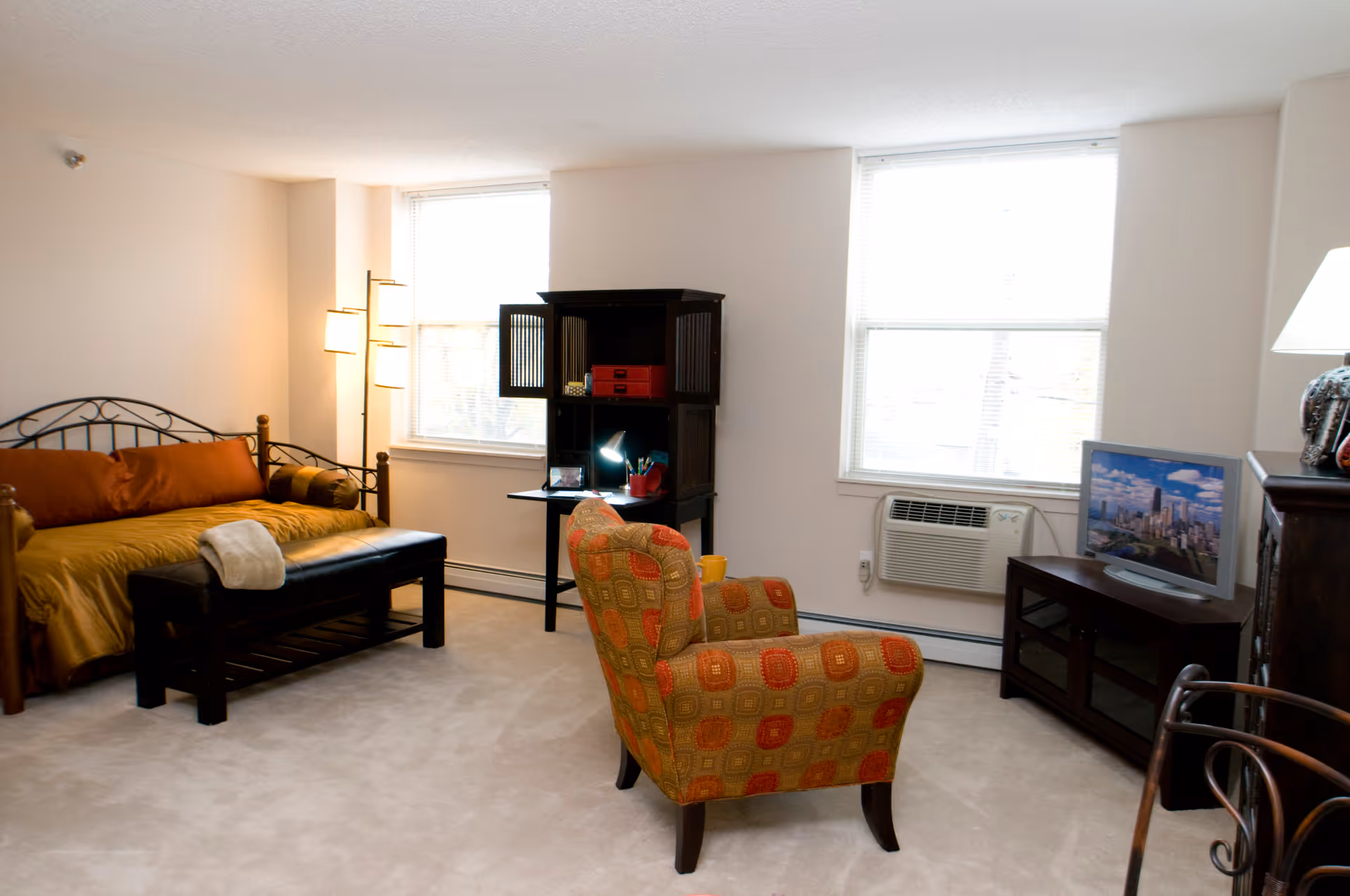 A cozy living room area in a senior living facility featuring a daybed with orange and brown pillows, a black bench with a white towel on it, a patterned armchair, a dark wooden cabinet with an open shelf and desk lamp, a TV on a dark wooden stand, two large windows with blinds, and a floor lamp in the corner.