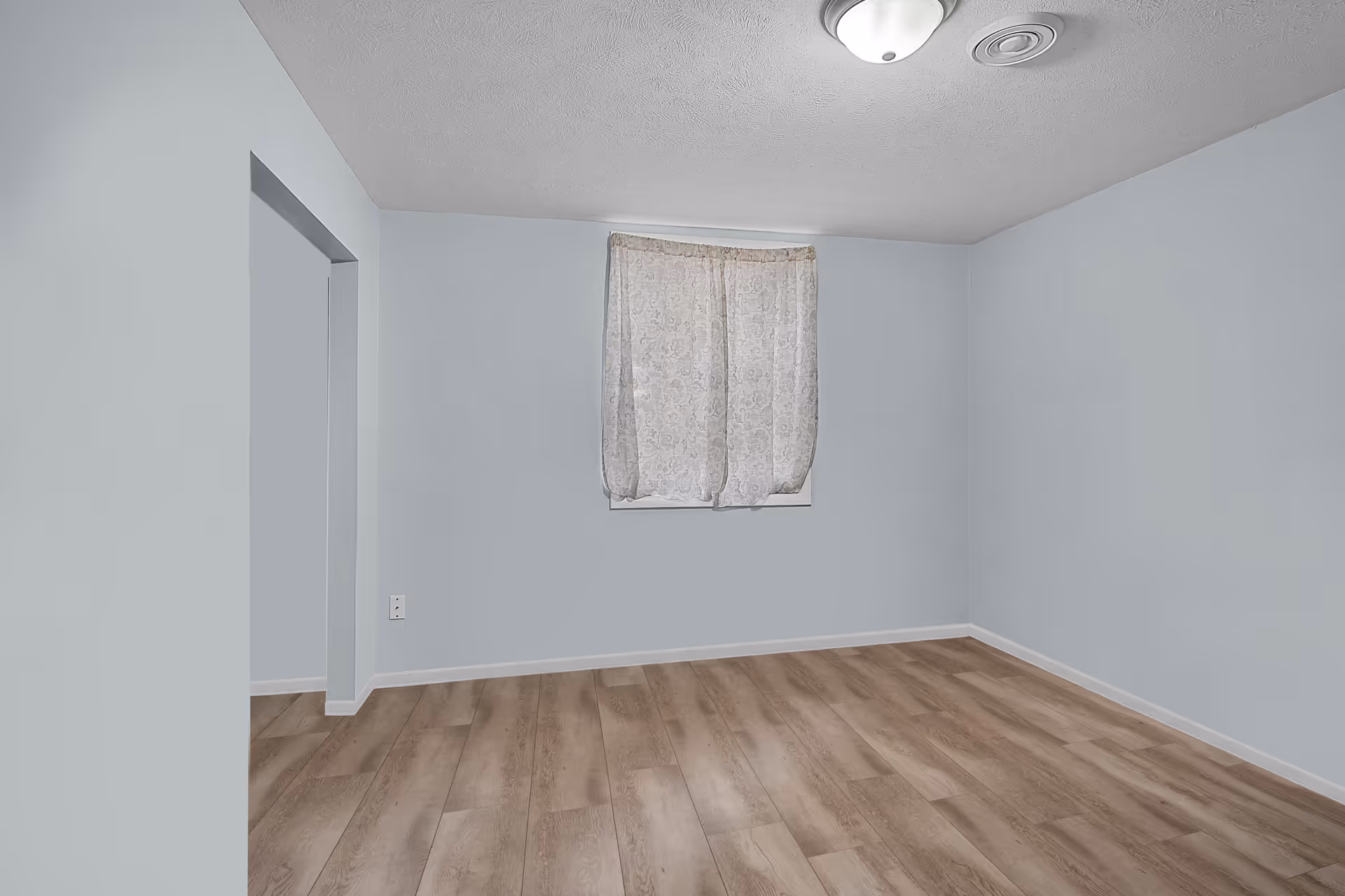 Empty small room with wood-look flooring, a single curtained window, and an overhead ceiling light.