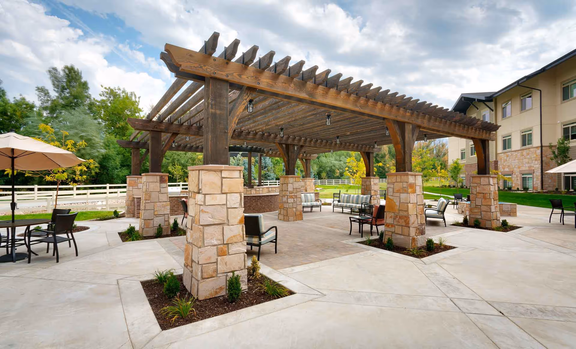 Covered outdoor patio with a large wooden pergola supported by stone pillars, surrounded by seating and a lawn near the senior living building.