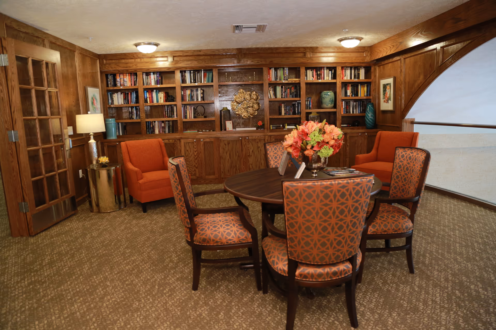 A cozy interior room with wood-paneled walls and built-in bookshelves filled with books and decorative items. There is a round wooden table in the center with a floral arrangement on top, surrounded by four patterned chairs. Two orange armchairs are placed against the wall near a small round side table with a lamp. A wooden door with glass panels is on the left side of the room.