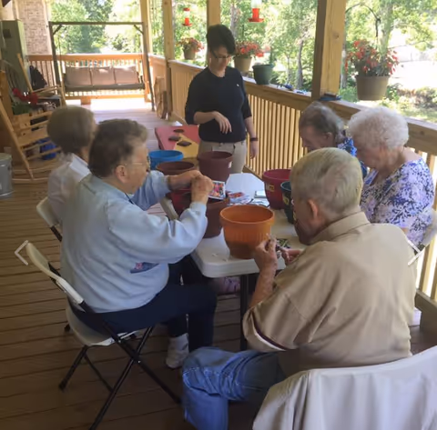 A group of elderly people sitting around a table on a wooden porch engaged in a planting activity with flower pots and soil. A caregiver or staff member stands nearby assisting them. The porch is decorated with potted plants and overlooks a green outdoor area.