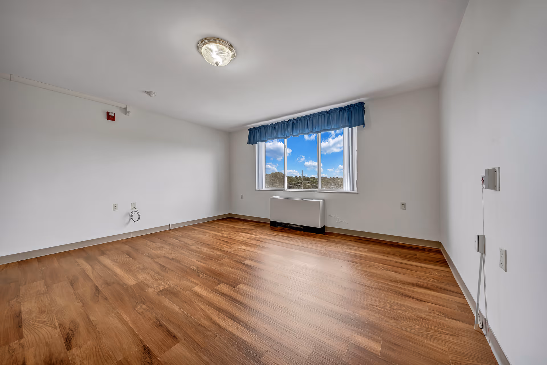 Empty room with wooden flooring, white walls, a ceiling light fixture, and a window with a blue valance showing a view of the sky and trees outside.