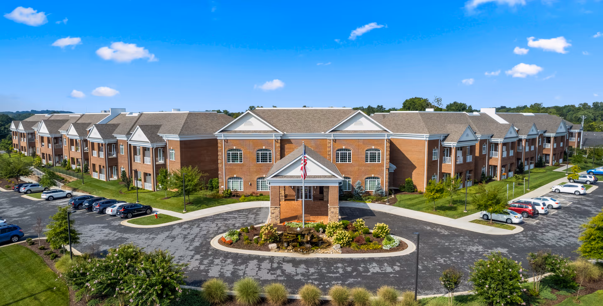 Exterior view of a large, two-story senior living facility building with a brick facade and white trim under a blue sky with scattered clouds. The building has multiple windows and a covered entrance with an American flag in front. There are landscaped gardens and parking lots with cars on both sides of the driveway leading to the entrance.