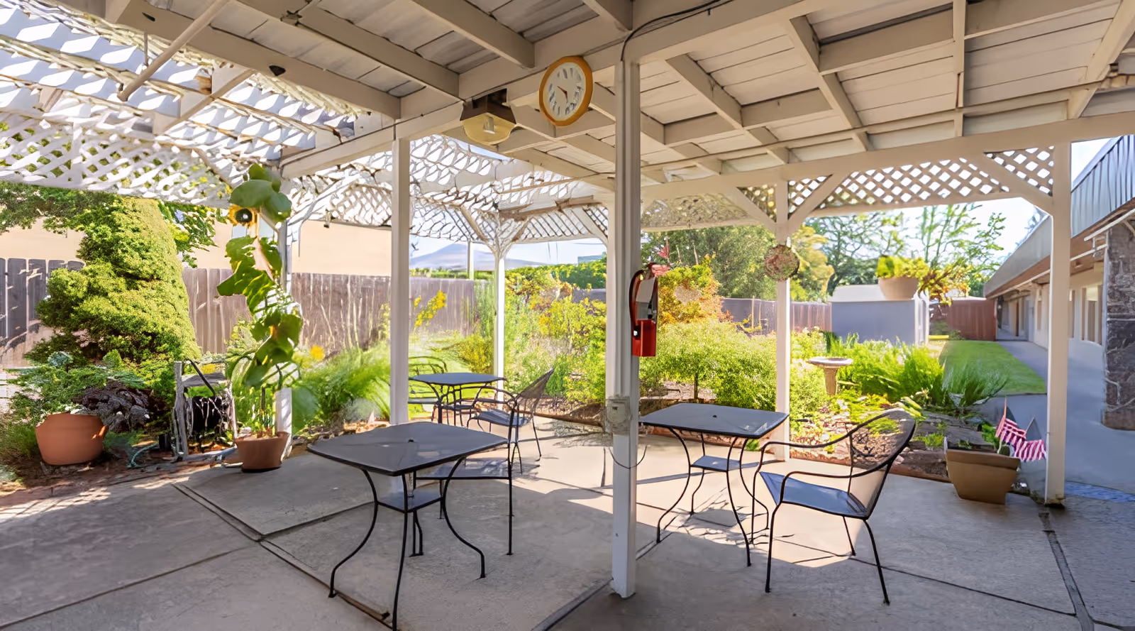 Outdoor covered patio area with metal tables and chairs, surrounded by lush greenery and potted plants. The patio has a white lattice roof and a clock hanging from the ceiling. There is a fire alarm mounted on a white pillar and a garden with various plants and a birdbath in the background.